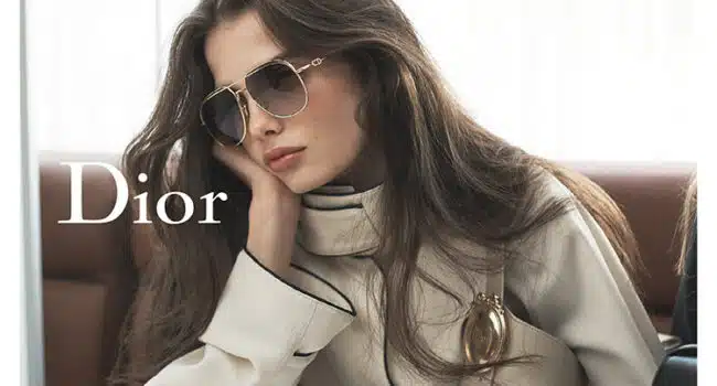 Woman wearing Dior sunglasses and a cream-colored high-neck blouse, resting her head on her hand while seated indoors. The Dior logo appears on the left side of the image.